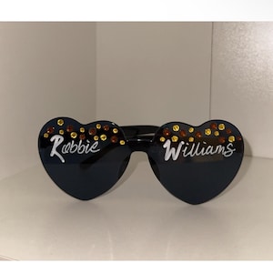 May include: Black heart-shaped sunglasses with the name "Robbie Williams" in white script. The top of each lens is decorated with gold and amber-coloured rhinestones. The sunglasses are displayed on a white surface.