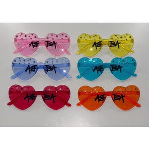 May include: Six pairs of heart-shaped sunglasses in various colours: pink, yellow, blue, turquoise, red, and orange. Each pair has the text "AB BA" and is decorated with rhinestones. A playful fashion accessory.