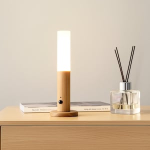 May include: A wooden bedside lamp with a cylindrical design, featuring a bright, rectangular light. The lamp has a wooden base and a small black button. A glass diffuser with reeds is also on the table.
