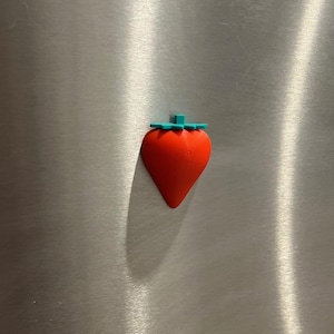 May include: A red strawberry-shaped magnet with a green stem, attached to a stainless steel surface. The magnet is a vibrant red color and has a smooth texture. The green stem adds a contrasting detail to the design.