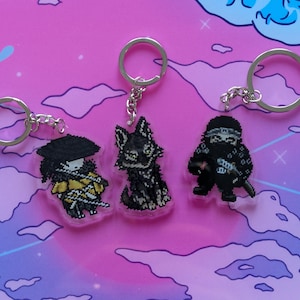 以下が含まれることがあります： ピクセルアートのキーホルダー3つ。黄色と黒のキャラクター、黒いオオカミ、忍者をそれぞれ描いています。各キーホルダーにはシルバーのキーリングとチェーンが付いています。背景は、青い波模様のピンクと紫のグラデーションです。