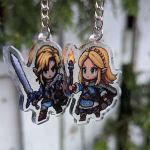 Könnte beinhalten: Zwei transparente Acryl-Schlüsselanhänger mit pixeligen Charakteren aus einem Videospiel. Einer zeigt einen Charakter mit Schwert, der andere mit einer Fackel. Beide Charaktere haben blondes Haar und tragen blaue und braune Outfits. Die Schlüsselanhänger hängen an silbernen Ketten.