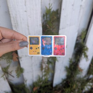May include: A sticker featuring three pixelated handheld game consoles in yellow, blue, and red. The sticker has a white border and is held against a white fence and green foliage background. The sticker is labeled "Heather Retro".