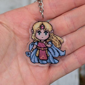 May include: A pixelated keychain featuring a character with blonde hair, a pink dress, and a blue cape. The keychain is attached to a silver chain. The character has a crown and green eyes. The design is reminiscent of a video game character.