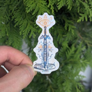 Op de afbeelding: Een holografische sticker met een zwaardontwerp en een gestileerd oogsymbool bovenaan. Het zwaard heeft blauwe en gouden accenten, met bloemen en geometrische patronen. De sticker wordt tegen een achtergrond van groen gebladerte gehouden.