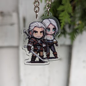 Puede incluir: Un llavero acrílico transparente con ilustraciones pixeladas de dos personajes con cabello blanco, un hombre y una mujer, de un videojuego de fantasía. El personaje masculino lleva una armadura marrón y sostiene una espada. El personaje femenino lleva una blusa blanca y pantalones oscuros.