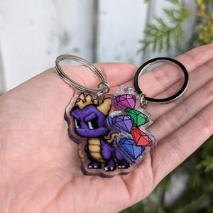 May include: A clear acrylic keyring featuring a pixelated purple dragon with a yellow crown and a cluster of colourful gems. The keyring has two silver key rings.
