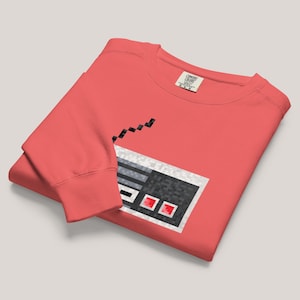 Könnte beinhalten: Korallfarbenes Sweatshirt mit einem pixeligen Videospiel-Controller-Design. Der Controller ist hauptsächlich grau und schwarz mit roten Knöpfen. Das Sweatshirt hat einen Rundhalsausschnitt und lange Ärmel, ideal für Freizeitkleidung.