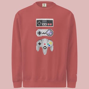 Könnte beinhalten: Ein ziegelrotes Sweatshirt mit pixeligen Illustrationen von drei Retro-Videospiel-Controllern: einem klassischen rechteckigen Controller, einem grauen Controller mit lila Akzenten und einem grauen N64-Controller mit bunten Knöpfen. Das Design ist mittig auf dem Sweatshirt platziert.