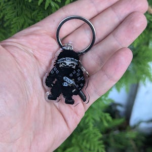 Könnte beinhalten: Ein klarer Acryl-Schlüsselanhänger mit einem pixeligen schwarzen Ninja mit einem silbernen Schlüsselring. Der Ninja trägt eine schwarze Kleidung mit einem weißen Gürtel und einem Schwert. Der Hintergrund ist verschwommenes grünes Laub.