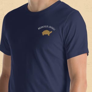 May include: Navy blue t-shirt with the text "BEHOLD, DOG!" above a gold turtle graphic. The short-sleeved shirt is made of a soft material and is suitable for casual wear.