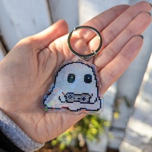 Könnte beinhalten: Ein pixeliger, geisterförmiger Schlüsselanhänger mit einem silbernen Schlüsselring. Der Geist ist weiß mit schwarzen Augen und einem Controller-Design. Der Schlüsselanhänger hat ein holografisches, irisierendes Finish, das mehrere Farben reflektiert.