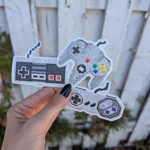 Op de afbeelding: Drie pixelachtige stickers van gamecontrollers. Eén is zwart-wit met een plusteken en rode knoppen. De andere twee zijn grijs met kleurrijke knoppen en pixelachtige ontwerpen. De stickers worden omhoog gehouden tegen een witte houten hek.