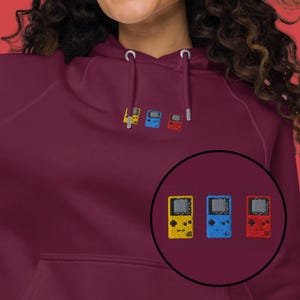 May include: Burgundy hoodie with embroidered retro handheld game console design. The design features three colorful consoles: yellow, blue, and red. The hoodie has a drawstring hood and a kangaroo pocket.
