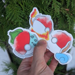 May include: A set of three die-cut stickers with a red, white, and blue design in a circular shape. One sticker has a green leaf and blue accents. The stickers are held against a backdrop of green foliage.