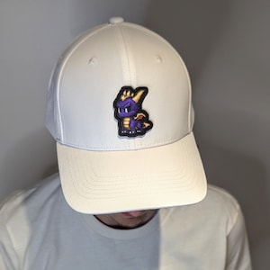 Pode incluir: Um boné de beisebol branco com um patch colorido de um dragão roxo com chifres e asas amarelas. O boné tem uma aba curva e uma coroa estruturada. O patch está centrado na frente.