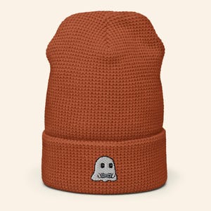 May include: A rust-coloured beanie hat with a textured knit pattern. The folded brim features a small embroidered ghost graphic. This warm hat is ideal for cooler weather and adds a touch of spooky fun to any outfit.