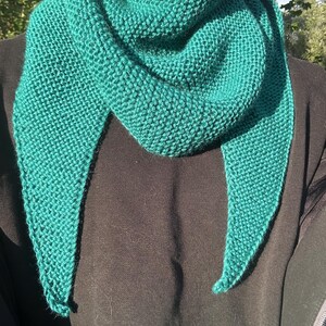May include: A triangular knit scarf in a vibrant teal color featuring a textured garter stitch pattern. The scarf is draped around the neck with two long, pointed ends hanging down over a solid black long-sleeved garment.