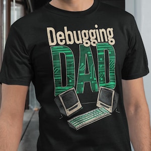 Op de afbeelding: Zwart T-shirt met de tekst "Debugging DAD" in groen en wit. Het ontwerp bevat een printplaatpatroon voor het woord "DAD" en computergraphics. Het shirt wordt gedragen door een persoon met een bril.