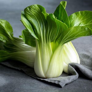 Puede incluir: Primer plano de bok choy fresco, una verdura de hoja verde, sobre un paño gris. Las hojas verdes vibrantes contrastan con los tallos blancos pálidos. La imagen destaca la textura y el color naturales del producto, ideal para listados culinarios.