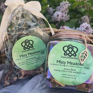 Puede incluir: Dos productos de aromaterapia: una bolsa para hervir a fuego lento y un tarro etiquetado "Misty Meadows" con eucalipto, hierba limón, cedro, romero y enebro. El tarro tiene una etiqueta de hoja de madera y la bolsa está atada con una cinta crema.