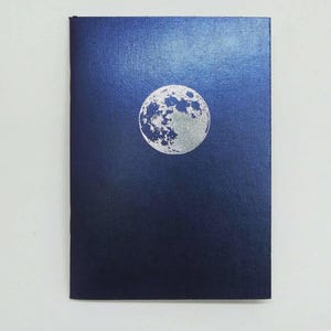 May include: A dark blue notebook with a silver moon design. The moon is centered on the cover and has detailed crater markings. The notebook has a textured surface and a rectangular shape.
