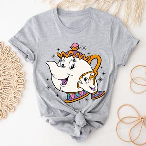 May include: A grey t-shirt with a cartoon image of Mrs. Potts and Chip from Beauty and the Beast. The design shows a white teapot with an orange handle and a small teacup, both smiling. The design is outlined in black with colourful accents.