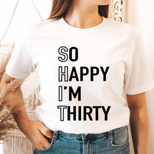May include: White t-shirt with the text "SO HAPPY I'M THIRTY" in black block letters. The shirt is a crew neck style and is being worn with blue jeans. The overall theme is a birthday celebration.