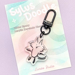 May include: Clear acrylic keychain charm with a black doodle of a cat-like creature. The charm is attached to a black metal clasp. The backing card has the text "Sylus Doodle" and "Glitter Epoxy Acrylic Keychain Charm".