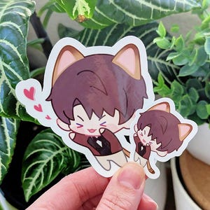 May include: Two stickers featuring a cartoon character with cat ears, brown hair, and a brown jacket. One sticker is larger than the other. The character is smiling and has hearts floating above them. The stickers are held in front of a green plant.