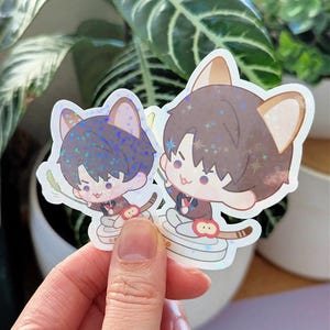 May include: Two stickers featuring anime-style characters with cat ears. One has blue hair, the other brown. Both are wearing brown jackets and holding a stick. The stickers have a holographic finish and are held in front of a plant.