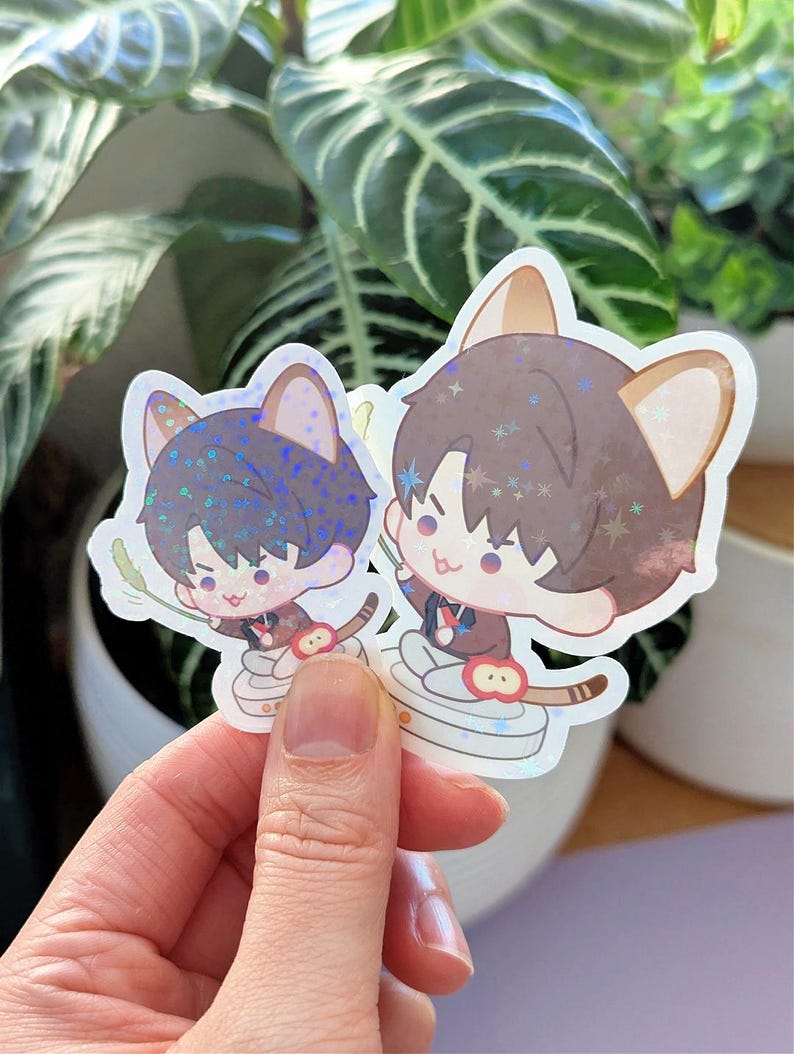 May include: Two stickers featuring anime-style characters with cat ears. One has blue hair, the other brown. Both are wearing brown jackets and holding a stick. The stickers have a holographic finish and are held in front of a plant.