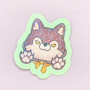 May include: A glittery sticker featuring a dark brown and cream-colored cartoon dog with yellow eyes and a yellow bow tie. The dog is depicted with its paws raised against a light green background, creating a playful and expressive character design.