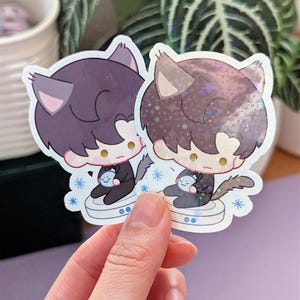 May include: Two cat-themed stickers. Each sticker features a cartoon cat character with dark hair, cat ears, and a small blue object. The stickers have a white border and are held in a hand.