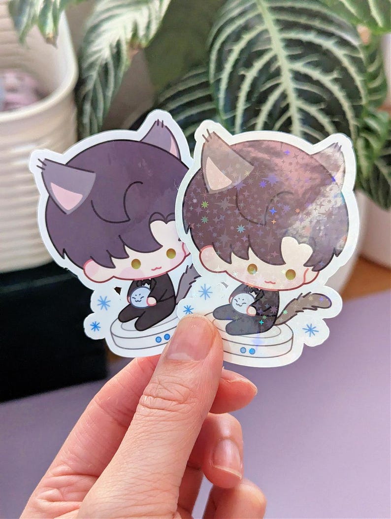 May include: Two cat-themed stickers. Each sticker features a cartoon cat character with dark hair, cat ears, and a small blue object. The stickers have a white border and are held in a hand.