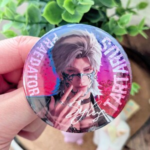 May include: A round button featuring a stylized illustration of a person with silver hair and a mask. The button has text that reads "TARTARUS" and "FRAEDATOR" in a circular pattern. The person's signature is also visible.