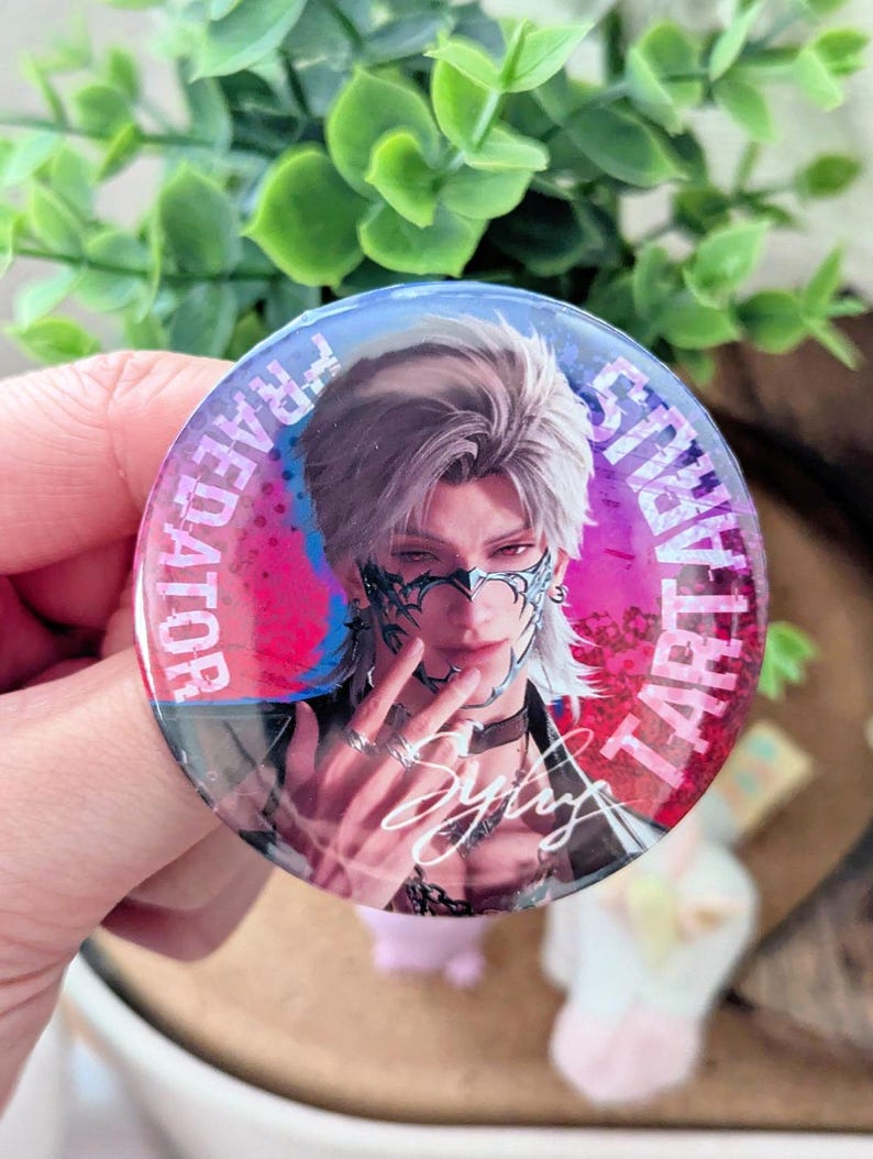 May include: A round button featuring a stylized illustration of a person with silver hair and a mask. The button has text that reads "TARTARUS" and "FRAEDATOR" in a circular pattern. The person's signature is also visible.
