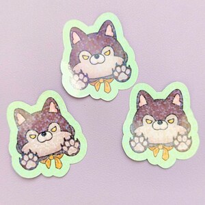 May include: Three die-cut stickers featuring a cartoon wolf character with dark purple fur, yellow eyes, and a yellow neck ribbon. The wolf has raised paws and a light green border. The surface of the stickers has a holographic glitter finish.