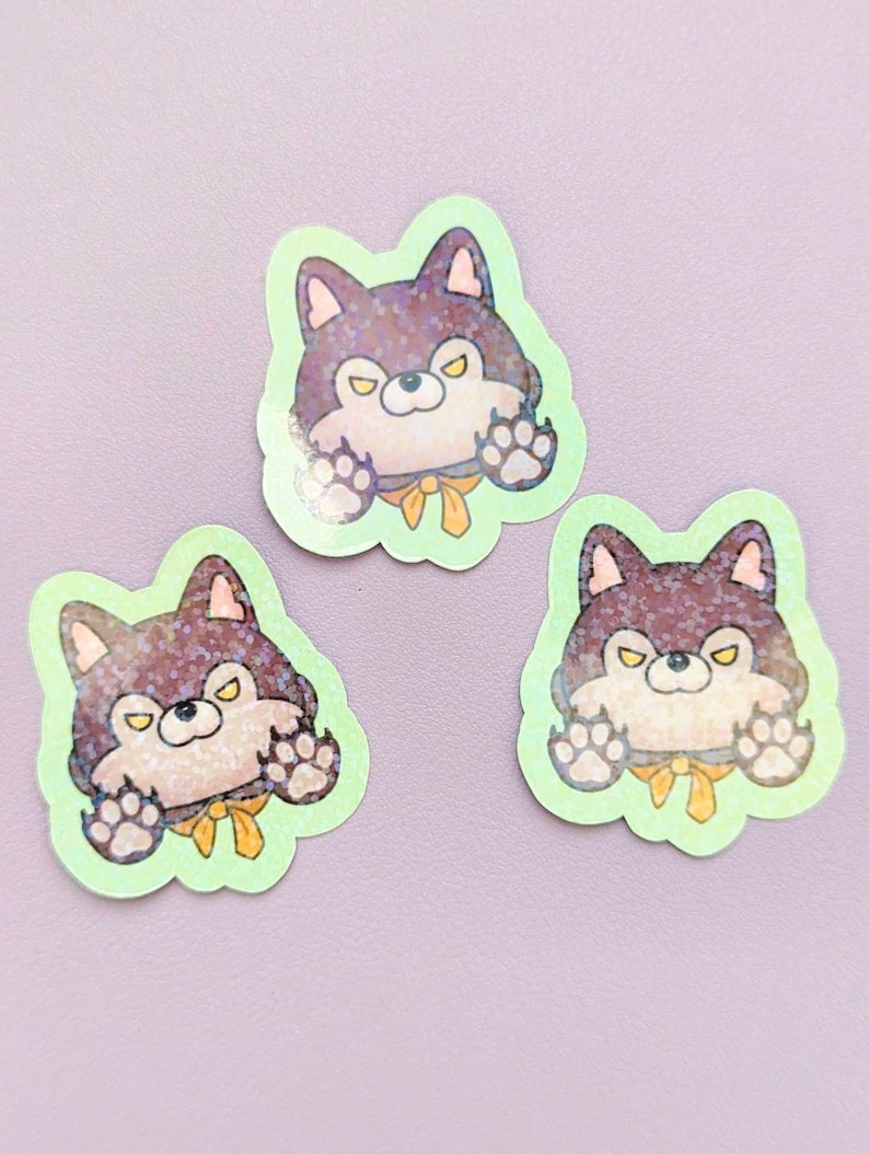 May include: Three die-cut stickers featuring a cartoon wolf character with dark purple fur, yellow eyes, and a yellow neck ribbon. The wolf has raised paws and a light green border. The surface of the stickers has a holographic glitter finish.