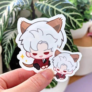 May include: Two stickers featuring a cartoon character with white hair, cat ears, and a red suit. One sticker is larger than the other. A yellow star is next to the larger sticker. The stickers are held by a hand.