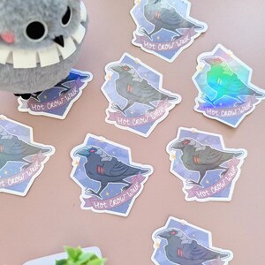 May include: A collection of stickers featuring a stylized crow design with the text "Hot Crow Walk" on a banner. The stickers have a holographic effect and are set against a light purple background. A plush bird toy is also visible.