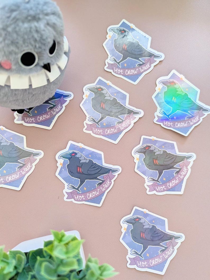 May include: A collection of stickers featuring a stylized crow design with the text "Hot Crow Walk" on a banner. The stickers have a holographic effect and are set against a light purple background. A plush bird toy is also visible.