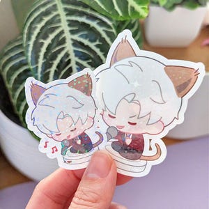 May include: Two holographic stickers featuring anime-style cat characters. The characters have white hair, brown ears, and are wearing suits. One is singing into a microphone, and musical notes are visible. The stickers are held in front of a plant.