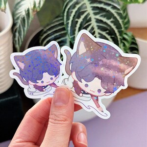 May include: Two cat-shaped stickers with anime-style illustrations. The stickers feature a purple and brown color scheme with glitter accents. The cat characters have cute expressions and are depicted in a dynamic pose, with a white outline.