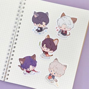 May include: A collection of five anime-style stickers featuring cat-eared characters in various poses. The stickers are on a grid-lined notebook page. Each character has different colored hair and outfits, with musical notes and other small details.
