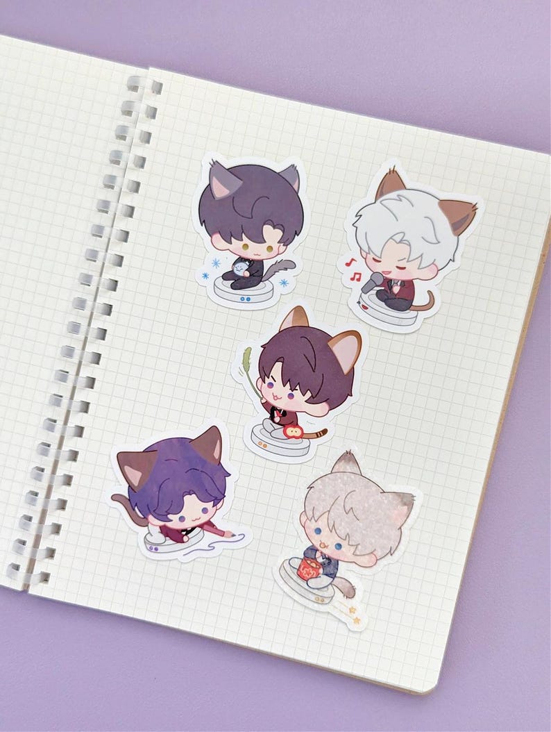 May include: A collection of five anime-style stickers featuring cat-eared characters in various poses. The stickers are on a grid-lined notebook page. Each character has different colored hair and outfits, with musical notes and other small details.