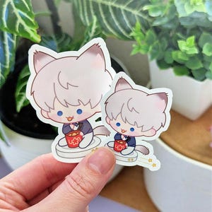 May include: Two stickers featuring a cartoon cat character with light gray fur, blue eyes, and a small red bucket. The cat is wearing a dark blue suit and sitting on a white platform. The stickers are held by a hand.