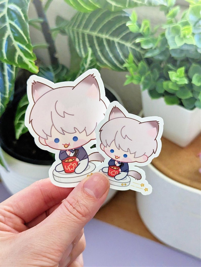 May include: Two stickers featuring a cartoon cat character with light gray fur, blue eyes, and a small red bucket. The cat is wearing a dark blue suit and sitting on a white platform. The stickers are held by a hand.