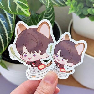 May include: Two stickers featuring a cartoon character with brown hair and cat ears, wearing a dark red outfit. The character is holding a green object. The stickers are on a white background.