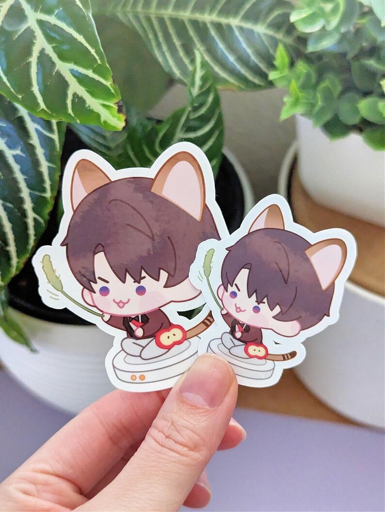 May include: Two stickers featuring a cartoon character with brown hair and cat ears, wearing a dark red outfit. The character is holding a green object. The stickers are on a white background.